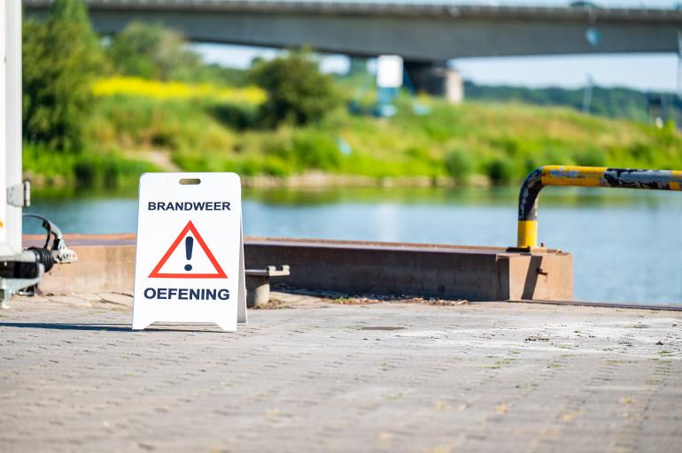 Bordje met tekst brandweer oefening en uitroepteken aan de rand van rivier de Maas