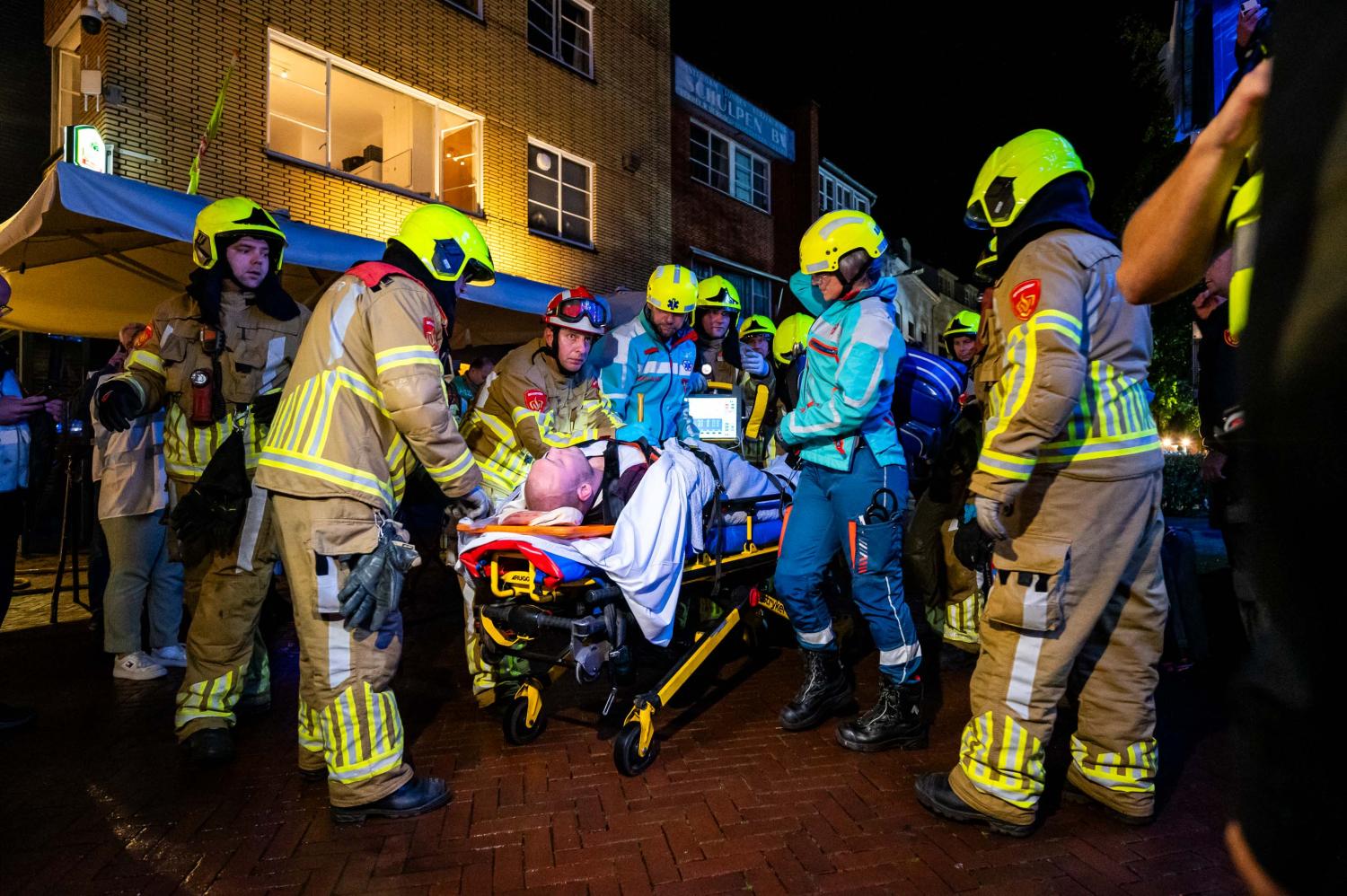 Man ligt op brancard. Hulpdiensten waaronder ambulance en brandweer staan eromheen.
